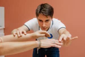 Praticien concentré en polo blanc examinant les mains d'un patient. Séance de kinésithérapie ou examen physique sur fond terracotta.