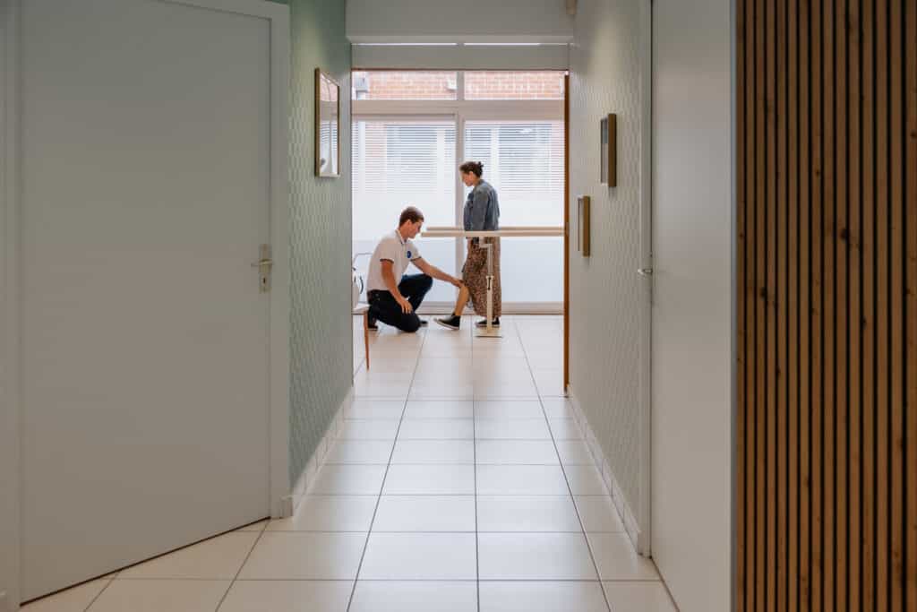 Kinésithérapeute en polo blanc examinant la jambe d'une patiente près de barres parallèles de rééducation dans un couloir lumineux.