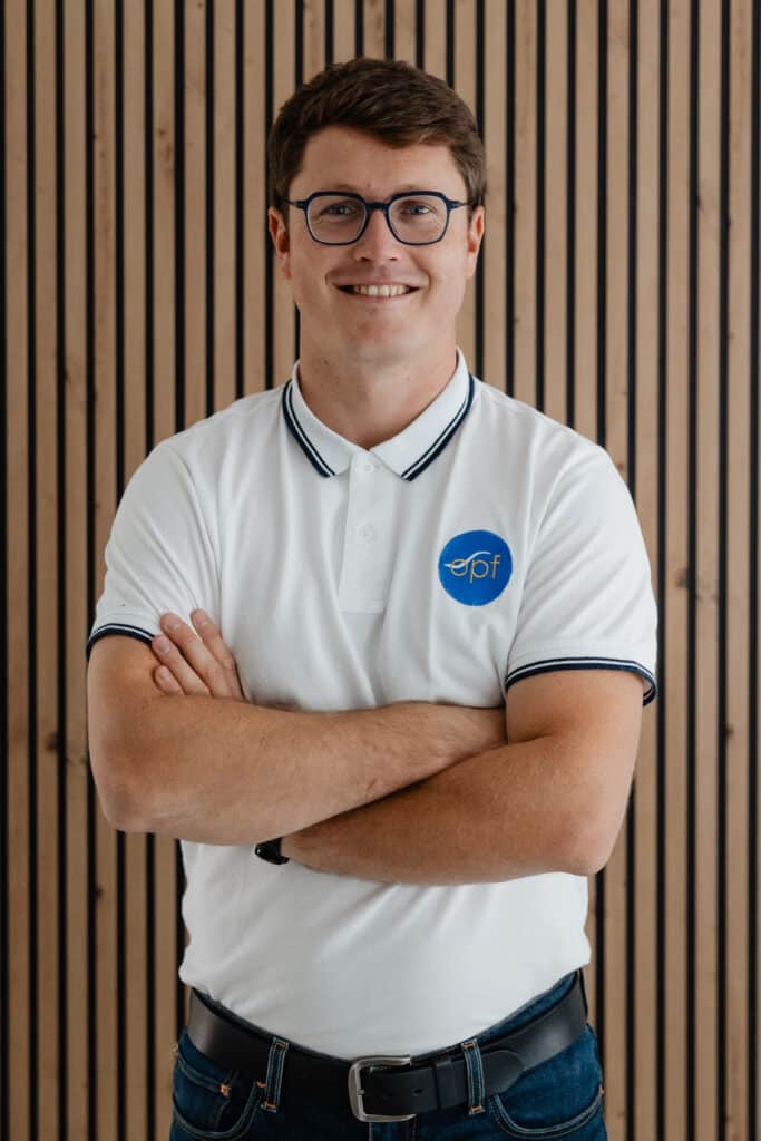 Homme souriant, polo blanc à rayures (logo EPF), lunettes, bras croisés. Portrait professionnel devant un fond acoustique moderne à lattes de bois.
