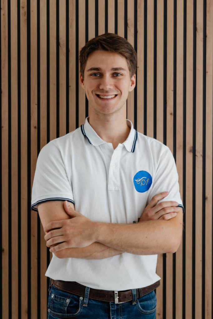 Jeune homme souriant en polo blanc avec le logo EPF brodé, les bras croisés, devant un mur à lattes de bois clair et noir.