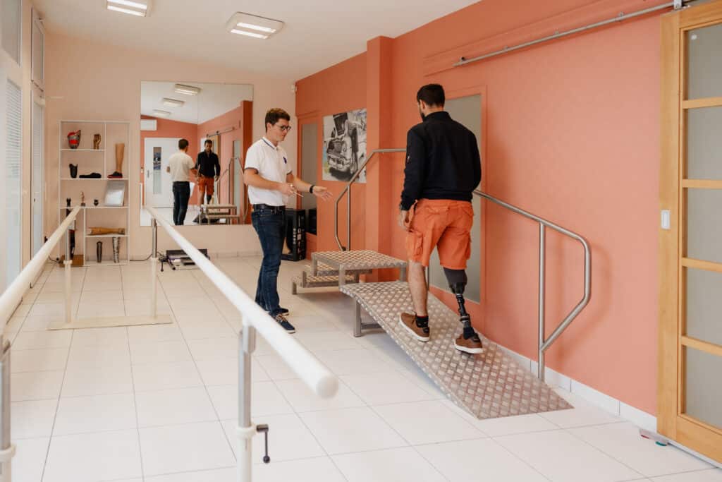 Patient avec prothèse s'entraînant à monter une rampe et des marches, guidé par un technicien. Centre de rééducation orthopédique.